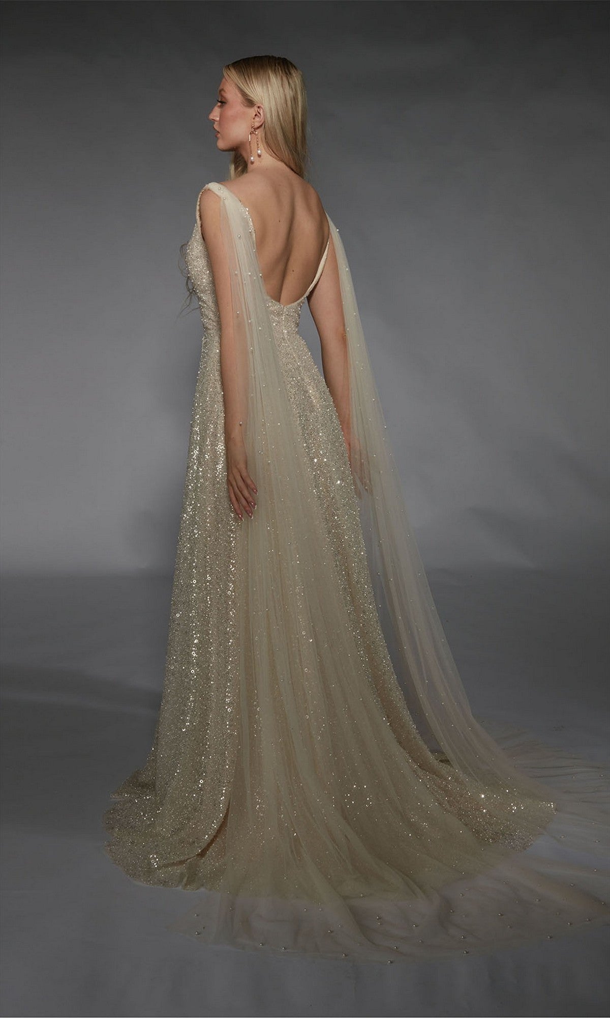 Woman wearing the Alyce 7134 long white dress with sequin embellishments, a deep back, and sheer cape-like panels, posing against a gray background.
